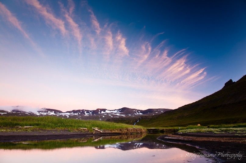 Peaceful cirrus clouds settle in streaks above a small river mouth. The distant mountains remain snowcapped to the end of summer.