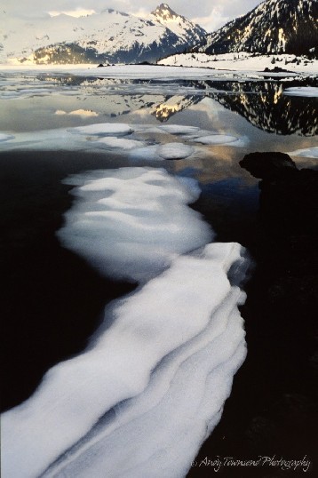 Layered patterns define ice floating on a lake with distant snowcapped mountains.