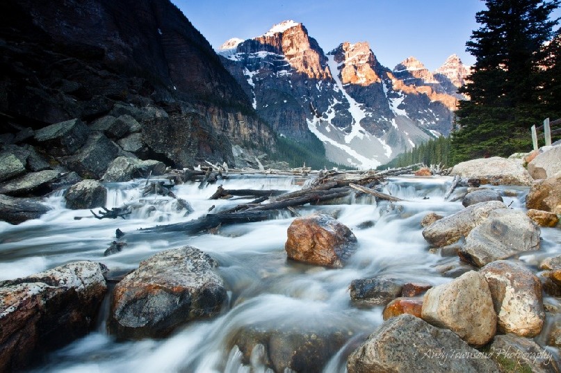 The Ten Peaks over Moraine Lake catch the morning sun while silken water flows over rocks and logs in the foreground.