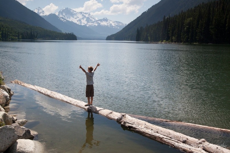 A boy balances on a log in a large lake with arms spread wide catching the sunshine with snow capped peaks in the distance.