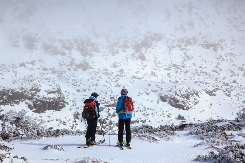 Two backcountry skiers pause on the ridge overlooking a distant cloud covered Rodway range.