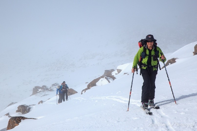 A female back-country skier skins up a track up the side of a ridge with two people in the distance.