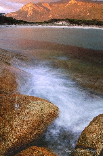 A long exposure shows swirling wave action against granite rocks with white sand and peaks of Mt Strezelecki beyond.