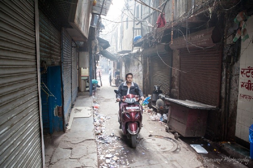 A young man rides a moped through an Old Dehli alleyway.