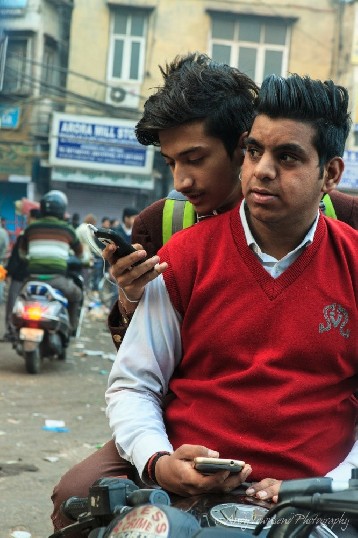 Two young Indian boys sit on their motorbike together while checking their mobile phones.