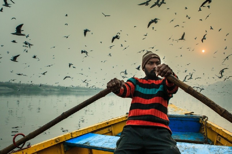 A man rows a wooden boat on the Yamuna river in Delhi with the sun breaking through the misty fog behind.