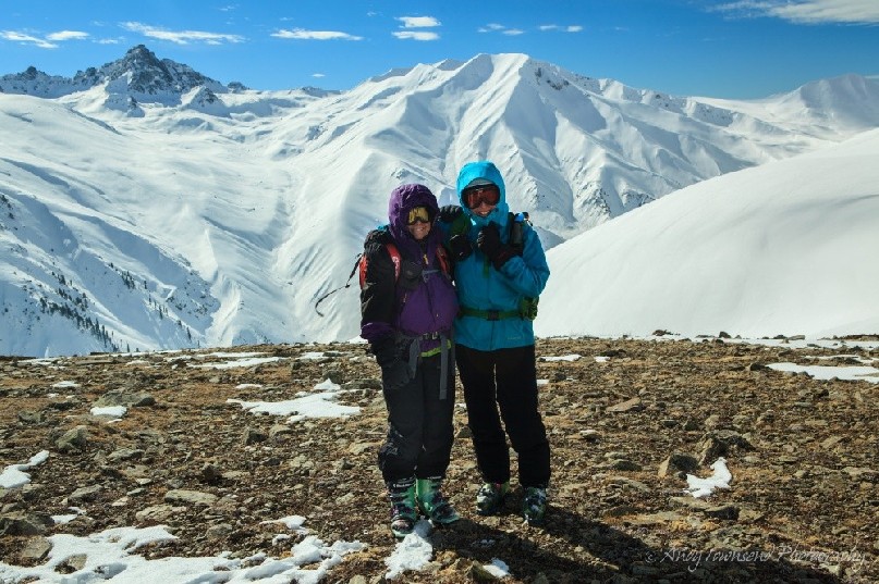 A couple of female skiers enjoy a hug on a bare patch of mountain top with a mountain range behind.