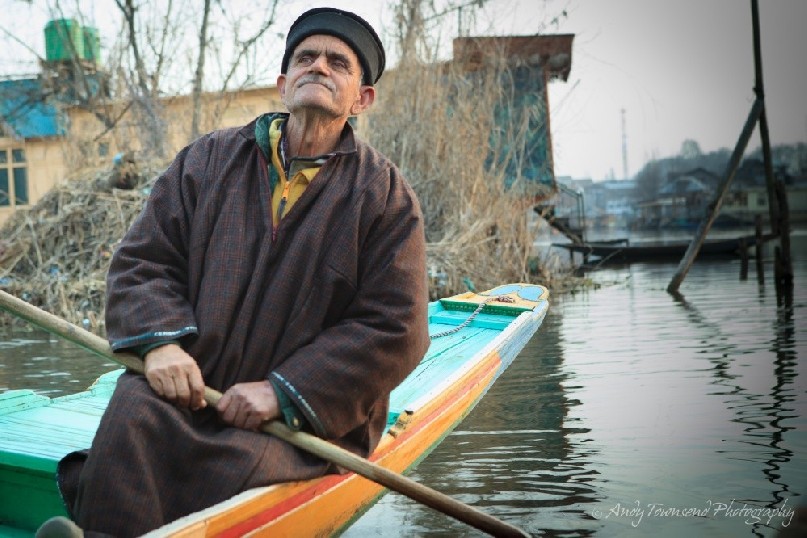 A man wearing a Phiran and beanie sits on the back of a Shikara looking into the distance.