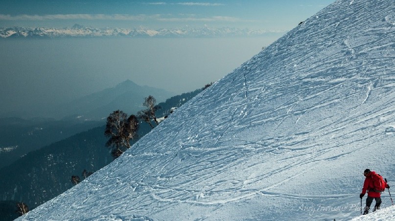 A ski guide pauses to check a slope for safety below a ski tracked slope. Beyond the slope are a fog-filled valley and mountain range.