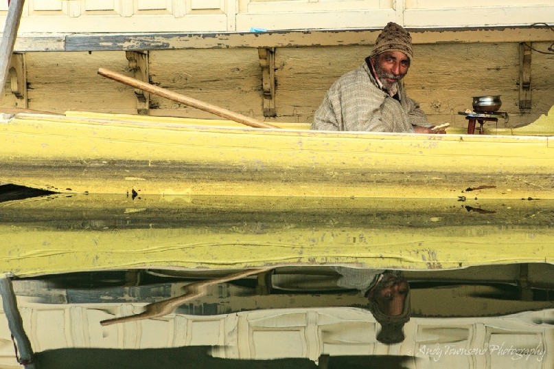 A man cooking in his Shikara wearing a Phiran and beanie, smiling with beard.