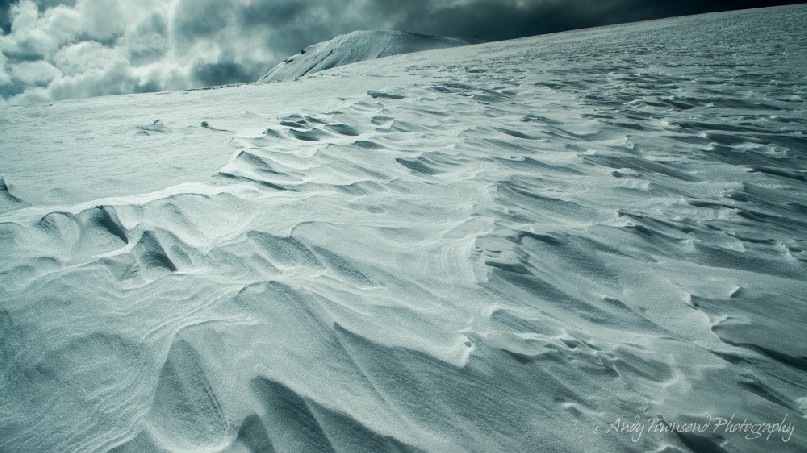 A line of wind-sculpted snow leads to a distant peak beyond.