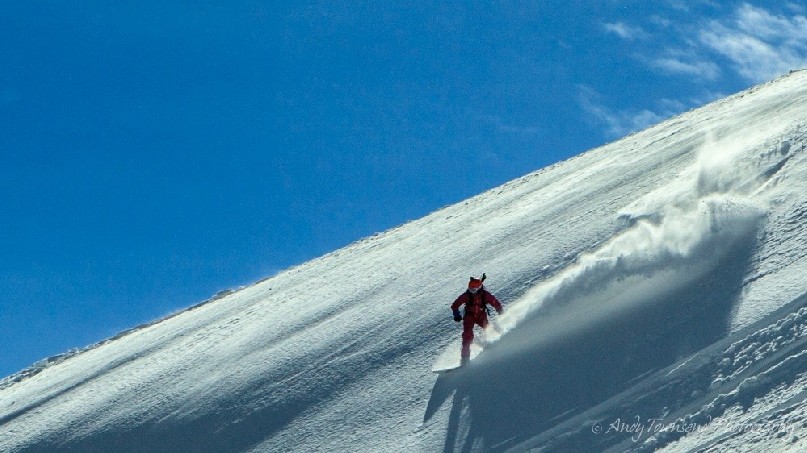 A snowboarder with plume of snow behind on a clear slope with clear sky.
