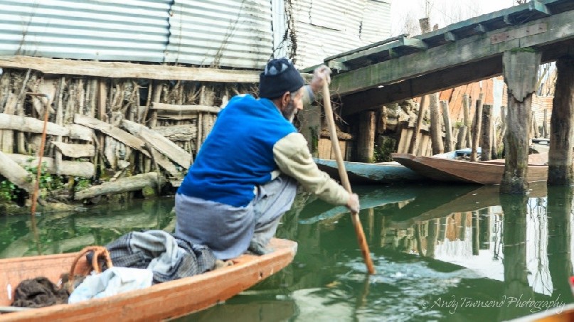 A man in mid-paddle navigates a narrow waterway off Dal Lake.