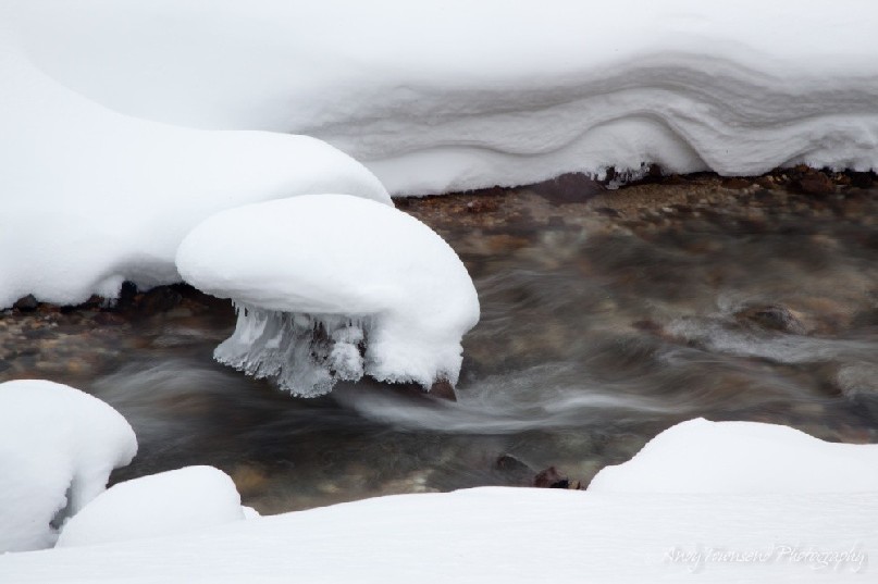 A rock adorned with layers of snow and ice sits in a stream surrounded by walls of snow