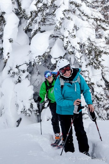 Two skiers pause midway up a skin track towards Furanodake mountain with snow-laden trees in the background.